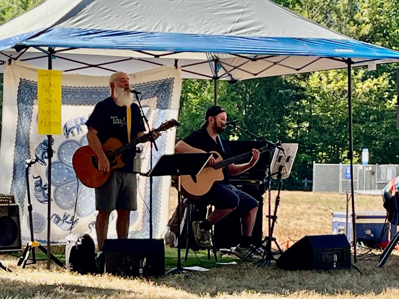 Carey Calhoun and Grant Stampfli playing an outdoor acoustic set under a pop-up canopy — Carey standing at the mic with his Taylor cutaway, Grant seated behind a music stand with an acoustic bass