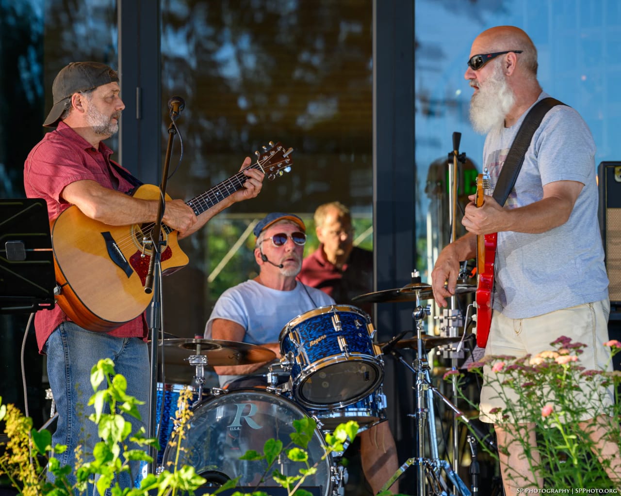 Phatt Ruby playing an outdoor stage in low evening sun — acoustic guitar at left, Mike Lynch behind the blue Rogers kit, Carey Calhoun on electric guitar at right