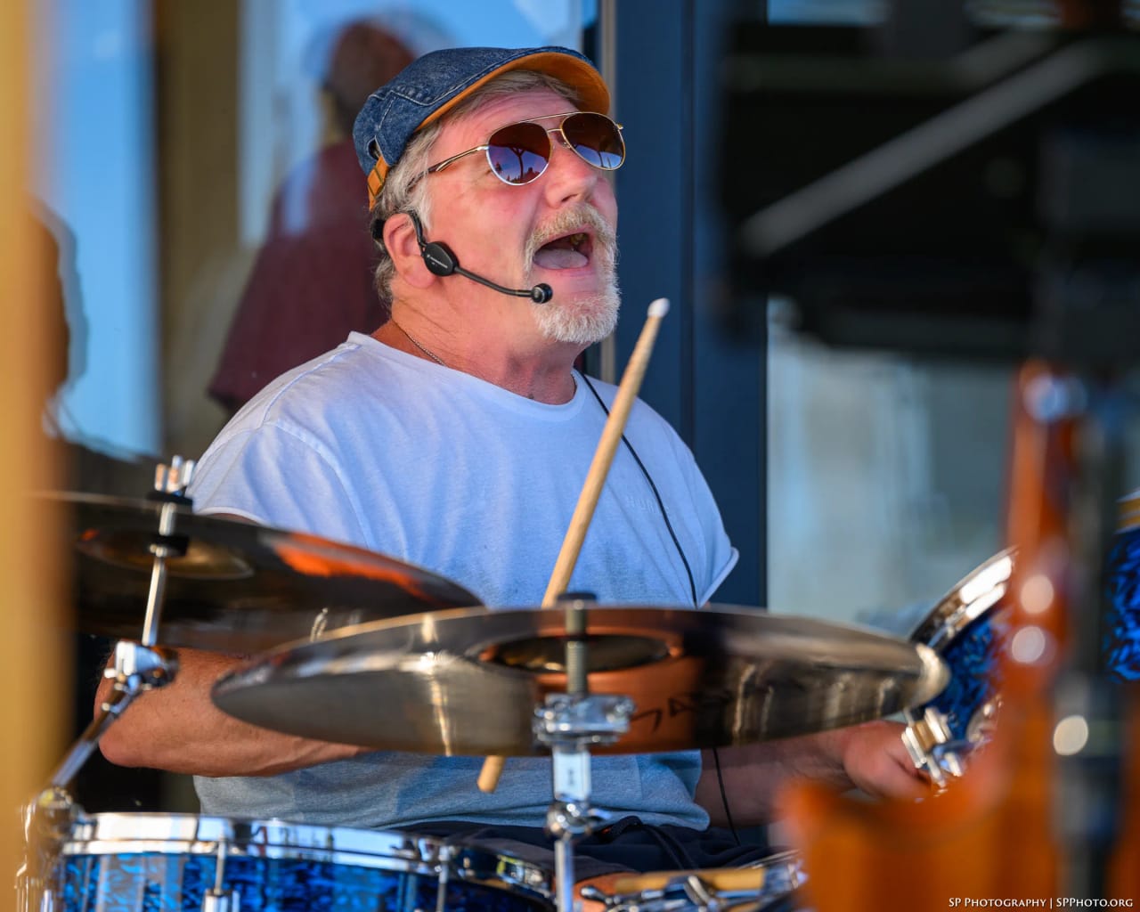 Mike Lynch singing into his headset mic behind the kit, stick raised, aviator sunglasses on