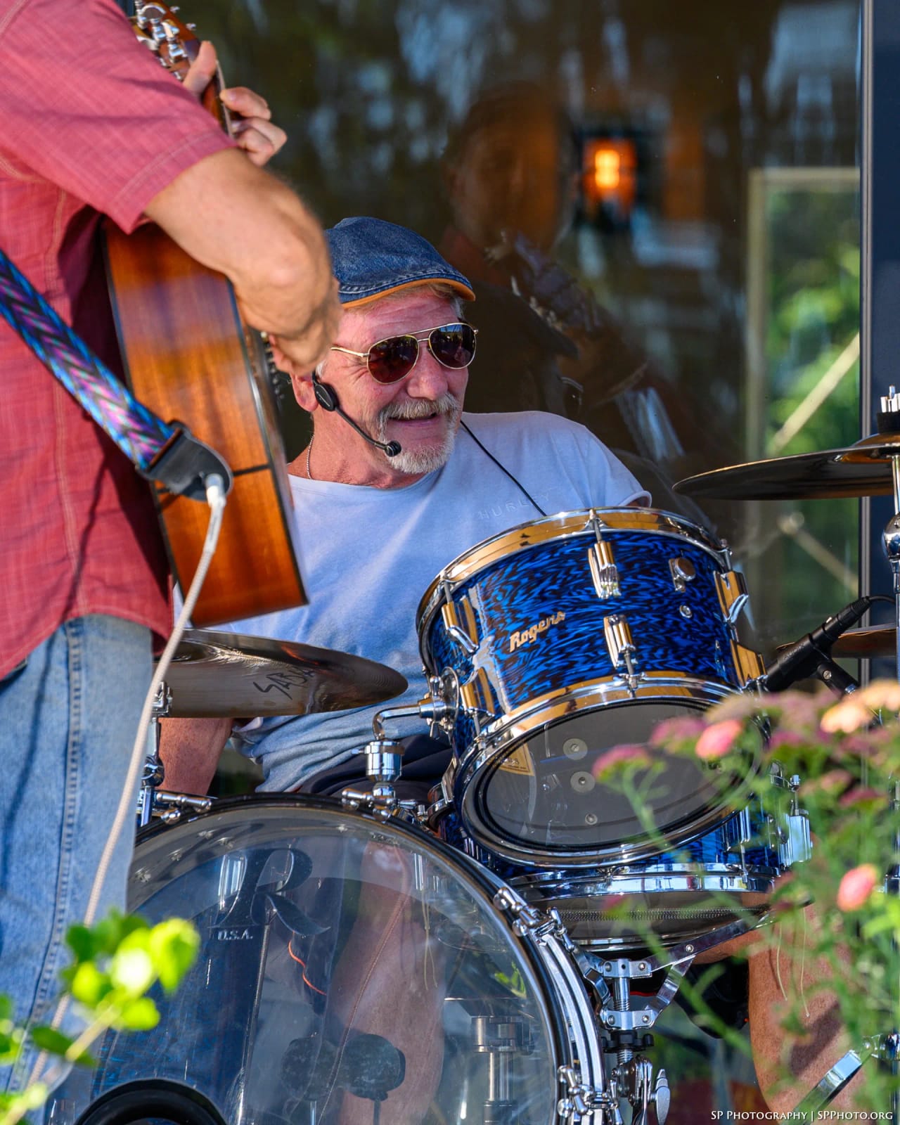 Mike Lynch grinning behind the blue Rogers kit, the neck of an acoustic guitar crossing the frame in front of him