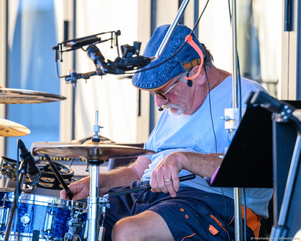 Mike Lynch in profile, head down over the kit mid-phrase, denim cap and in-ear monitor