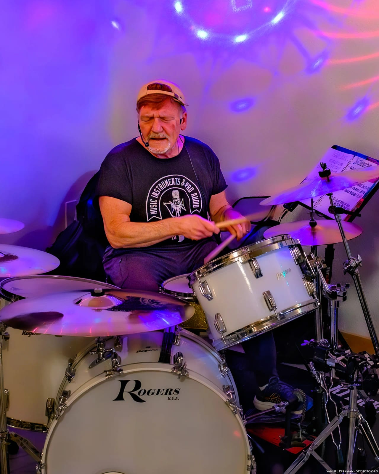 Mike Lynch behind his white Rogers kit, sticks mid-stroke, purple stage lights washing the wall behind him